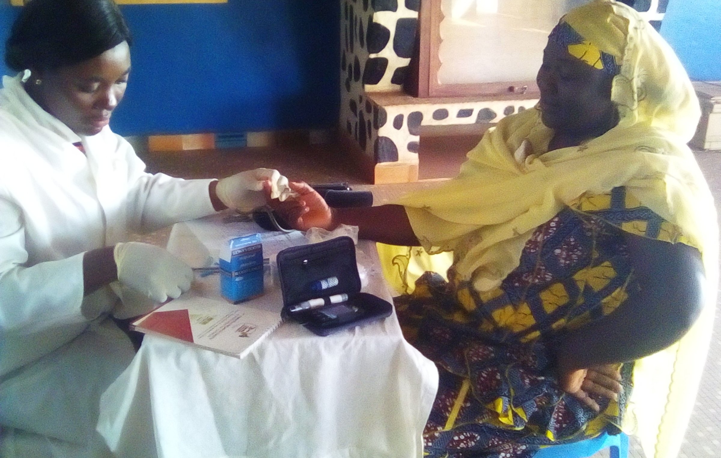 L'image montre une scène dans une salle de consultation. À gauche, une professionnelle de la santé, vêtue d'un manteau blanc et de gants, effectue un test sur le bras d'une patiente. La femme qui reçoit le soin porte un turban jaune et une robe aux motifs colorés. Elle semble concentrée et un peu nerveuse. Sur la table devant elles, on peut voir des documents et des instruments médicaux, comme un petit appareil de mesure. Les murs de la pièce sont peints en bleu, et le sol est en béton. L'environnement est calme, et l'atmosphère est professionnelle, axée sur le soin et le soutien.