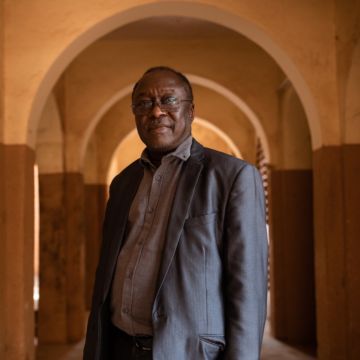 L'image montre un homme debout dans un espace intérieur, caractérisé par de grandes arcades. Les murs sont peints de couleurs chaudes, avec des tons ocre et beige. L'homme porte une veste sombre et une chemise grise, et il a l'air confiant. Son expression est sereine et il regarde légèrement vers la droite. L'arrière-plan présente des arches qui créent une atmosphère de profondeur et de perspective. La lumière semble douce et diffuse, ajoutant une ambiance chaleureuse à la scène.