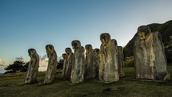 The image features a series of large stone sculptures arranged in a row, resembling human figures. These sculptures have distinct, elongated shapes and facial features, with some appearing to look downward. The background includes a grassy area and a hillside, with trees visible nearby. The sky above is clear, transitioning from blue to lighter tones as it approaches the horizon, suggesting either dawn or dusk. The overall atmosphere gives a sense of mystery and historical significance to the figures.