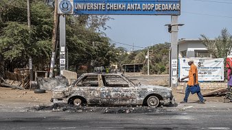 L'image montre l'entrée de l'Université Cheikh Anta Diop de Dakar, identifiable par le panneau au-dessus. En premier plan, une voiture calcinée est garée sur la route, avec des traces de brûlures et des dégâts visibles, indiquant qu'elle a été abandonnée après un incendie. En arrière-plan, on peut apercevoir des arbres et des structures, mais l'atmosphère semble marquée par un sentiment de désolation. Un piéton, portant une sacoche, marche sur le trottoir, ajoutant un contraste entre l'activité humaine et l'état dégradé des lieux.