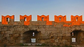 L'image montre un mur de fortification ancien, construit en pierres de taille. La lumière dorée du soleil se reflète sur la pierre, créant un effet chaleureux et pittoresque. Au sommet du mur, on peut voir des créneaux, typiques des châteaux, qui forment des formes découpées. Le mur présente des arches et des ouvertures qui laissent entrevoir un espace en arrière-plan, probablement une vue sur la mer ou un paysage naturel. Le sol est recouvert de gravier, ajoutant une texture à l'environnement. L'ensemble dégage une atmosphère historique et paisible.