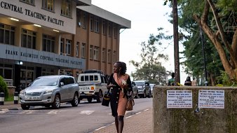 Dans cette image, on voit une scène urbaine près d'un bâtiment nommé "Frank Kalimuzo Central Teaching Facility". La structure est moderne avec plusieurs fenêtres et un design clair. Sur le trottoir, une femme vêtue d'une robe rose marche, portant une sacoche. À l'arrière-plan, on aperçoit quelques véhicules, dont une voiture blanche, et des arbres qui apportent une touche de verdure à l'environnement. Des panneaux sur un mur en béton à droite attirent l'attention avec des avertissements écrits en grandes lettres. L'ambiance est calme, suggérant un moment de transition entre la journée et le soir.