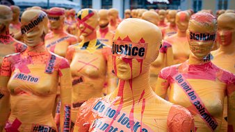 L'image montre un vaste rassemblement de silhouettes humaines couvertes de plastique coloré. Chaque silhouette est peinte en orange vif et ornée de mots et de slogans en noir et en rouge, évoquant des thèmes sociaux et des mouvements de droits civiques comme #MeToo et la justice. Certains mots sont barrés ou soulignés pour accentuer leur impact. L'atmosphère dégage un sentiment fort d'engagement et de protestation, avec ces figures anonymes qui semblent former un mur de revendication. L'ensemble crée une installation artistique saisissante, incitant à la réflexion sur des enjeux sociaux importants.