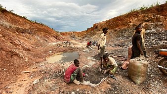 L'image montre une scène rurale d'exploitation minière. On y voit un paysage marqué par des fouilles, avec un sol rouillé et des montagnes de terre séparées. Au premier plan, plusieurs personnes, principalement des hommes et des jeunes garçons, sont en train de travailler. Certains sont accroupis, tamisant des pierres ou ramassant des minéraux, tandis que d'autres semblent transporter des sacs pleins de matériaux. À gauche, il y a un petit plan d'eau, qui est trouble, entouré de débris miniers. Le ciel est nuageux, ajoutant une ambiance sombre à la scène, évoquant un travail difficile et épuisant.