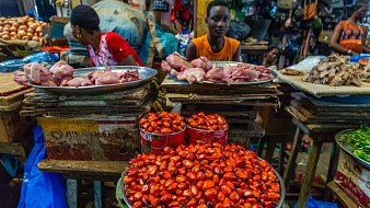 L'image montre un marché animé, où des étals en bois sont chargés de différents aliments. Au premier plan, un grand plat en métal contient des petites noix rouge vif, à côté d'autres aliments. À l'arrière-plan, des morceaux de viande sont disposés sur des plateaux, et on peut voir des vendeurs et des clients s'affairer autour. Les couleurs sont vives, avec des nuances de rouge, de marron et de vert. L'atmosphère est dynamique, remplie de sons de discussions et d'échanges.