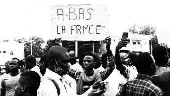L'image montre une scène de manifestation. Au centre, une personne se tient debout, brandissant une grande pancarte sur laquelle est écrit "À BAS LA FRANCE". Cette phrase semble exprimer un sentiment de mécontentement. Autour de cette personne, un groupe d'hommes et de femmes montre diverses expressions, allant de la détermination à la colère. Certains tiennent des téléphones portables, prenant des photos ou des vidéos de la manifestation. L'arrière-plan est flou, mais on peut distinguer des bâtiments et des arbres. L'atmosphère générale dégage une forte émotion et un engagement collectif sur un sujet qui préoccupe les manifestants.