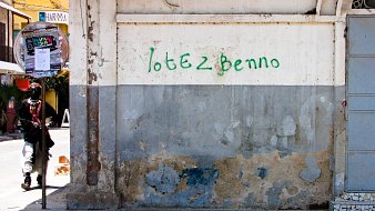 L'image montre un mur d'un bâtiment, principalement de couleur beige, avec des marques d'usure et de décoloration, témoignant de son ancienneté. Une inscription verte, "Votez Benno", est peinte sur le mur. À gauche, on aperçoit la silhouette d'un individu habillé de manière décontractée, qui semble marcher. Au fond, il y a une ambiance animée avec des éléments urbains tels que des affiches et des tables à l'extérieur de cafés. L'ensemble dégage une atmosphère de quartier vivant.