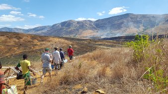 La scène dépeint un paysage rural habité par des personnes marchant sur un terrain partiellement brûlé. Au premier plan, plusieurs individus, habillés de vêtements décontractés, avancent avec des outils ou des équipements à la main. L'environnement est dominé par des herbes sèches et des terres noircies, traces d'un feu récent. À l'arrière-plan, on aperçoit des collines et des montagnes qui se dressent majestueusement sous un ciel bleu parsemé de nuages blancs. L'atmosphère est celle d'une randonnée dans la nature, mêlant effort humain et les conséquences d'une activité agricole ou un incendie. L'ensemble évoque une interaction entre l'homme et son environnement, avec une certaine gravité due à la destruction visible.