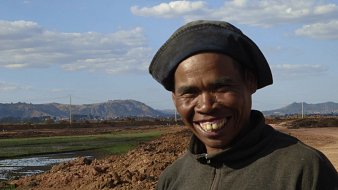 L'image montre un homme souriant se tenant devant un paysage rural. Il porte un chapeau noir et un vêtement sombre. En arrière-plan, on peut voir des collines et un ciel parsemé de nuages blancs. Le sol est terreux, et il y a des zones d'eau, probablement des rizières. L'expression de l'homme dégage de la joie et de la chaleur, ajoutant une touche positive à cette scène naturelle.