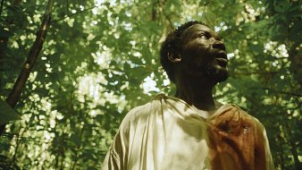 L'image montre un homme debout dans une jungle luxuriante. Il porte une tunique légère, dont une partie est tachée de rouge, suggérant un événement émotionnel ou tragique. Le visage de l'homme exprime la surprise ou l'inquiétude, avec des traits marqués par la lumière douce qui filtre à travers le feuillage dense. Les feuilles vertes et les ombres créent une atmosphère à la fois mystérieuse et intense, plongeant le spectateur dans l'ambiance d'une nature vibrante et sauvage. On peut imaginer les sons de la jungle, comme le chant des oiseaux et le bruissement des feuilles, qui entourent l'homme.