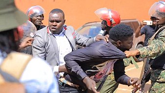 L'image montre une scène tendue sur une rue. On peut voir des policiers en uniforme, portant des casques rouges, qui semblent intervenir auprès d'un homme. Ce dernier est tiré en arrière par un autre homme qui essaie de le protéger. L'atmosphère est chaotique, avec des tensions visibles entre les forces de l'ordre et la personne qu'ils maîtrisent. En arrière-plan, il y a quelques personnes observant la situation et des véhicules garés. Le paysage évoque une ambiance de manifestation ou d'émeute.