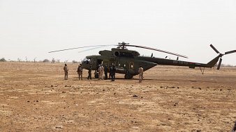 L'image montre un hélicoptère posé sur un terrain désertique, avec un sol caillouteux et sec. Autour de l'hélicoptère, plusieurs personnes sont rassemblées, certaines en uniforme militaire. La scène semble se dérouler dans une région aride, avec peu de végétation en arrière-plan.