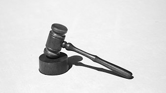L'image présente un maillet de juge, souvent appelé "gavel". Il est en métal noir, avec une partie cylindrique plus épaisse à sa base et un long manche. Le maillet repose sur une surface claire, ce qui crée un contraste marqué. La lumière éclaire l'objet, projetant une ombre sur le côté. Le maillet est couramment associé au monde de la justice et des tribunaux, symbolisant l'autorité et la prise de décision.