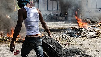 L'image montre une scène intense de protestation. Un homme, portant un débardeur blanc et un bandana sur le visage pour se protéger, est vu en arrière-plan. Il transporte un pneu que semble-t-il il va utiliser pour le jeter dans un feu. Autour de lui, on aperçoit de la fumée épaisse s'élevant dans l'air, résultant de plusieurs feux allumés sur la route. Le sol est jonché de débris, et l'atmosphère est chargée de tension, évoquant un sentiment de chaos et de lutte. Les flammes crépitent et laissent émaner une lumière orange. En arrière-plan, on peut distinguer des silhouettes de personnes, probablement d'autres manifestants.