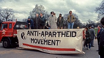 Cette image représente un rassemblement pour le mouvement anti-apartheid. On voit une scène sur un camion, avec plusieurs personnes qui parlent à un microphone. La bannière au premier plan affiche le texte "ANTI-APARTHEID MOVEMENT" en lettres majuscules, souligné par une bande rouge. L'environnement est urbain, avec des arbres dénudés en arrière-plan et un ciel nuageux, ce qui crée une atmosphère sérieuse et engagée. Les participants semblent passionnés, témoignant de l'importance de la lutte pour l'égalité et les droits civiques.