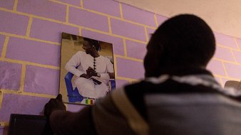 Dans cette image, un homme est en train de fixer un portrait au mur. Il se tient de dos, face à un mur peint en violet aux briques jaunes. Le portrait montre une personne assise, vêtue d'une tenue traditionnelle, probablement sénégalaise, avec un fond flou. L'homme qui fixe le portrait semble concentré sur sa tâche, ajoutant une touche personnelle à l'espace où il se trouve.