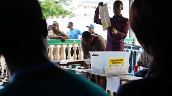 L'image montre une scène d'élections parlementaires. Au centre, un homme se tient debout, levant une feuille de papier, probablement un bulletin de vote, pour montrer aux personnes présentes. Il s'habille d'une chemise de couleur sombre et semble expliquer quelque chose de manière engageante. En arrière-plan, plusieurs personnes sont rassemblées, certaines observant attentivement tandis que d'autres attendent, adossées à une balustrade. On devine une atmosphère de concentration et d'anticipation, typique d'un moment électoral. Sur une table, une boîte transparente étiquetée "ELECTIONS PARLEMENTAIRES" est visible, contenant probablement des bulletins de vote. 