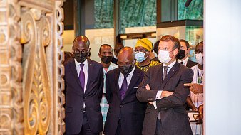 L'image montre un groupe de personnes, dont trois hommes, en train d'observer un objet d'art ou une exposition. Les hommes sont habillés de costumes formels, avec deux en costumes sombres et un en costume gris avec une cravate. Ils affichent des expressions de concentration et d'intérêt. À l'arrière-plan, on perçoit des éléments culturels, probablement des œuvres d'art ou des artifacts, créant une ambiance muséale. Les visiteurs portent tous des masques, ce qui indique qu'ils se trouvent dans un contexte où cela est nécessaire, peut-être en raison de préoccupations sanitaires. Le cadre est bien éclairé, mettant en valeur l'importance de l'exposition.