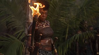 L'image montre une femme debout au milieu d'une luxuriante végétation tropicale, avec des palmiers en arrière-plan. Elle tient une torche enflammée dans sa main droite, créant une lumière chaleureuse qui éclaire son visage déterminé. Elle porte un haut traditionnel en cuir et une jupe décorée de motifs. Des bracelets dorés ornent ses bras, ajoutant une touche d'élégance à son apparence. Son expression est forte et intense, reflétant à la fois courage et détermination. L'atmosphère de l'image évoque une ambiance de puissance et de leadership.