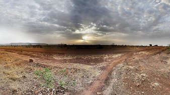 Imaginez un vaste paysage ouvert, dominé par des tons chauds de terre et des nuances de beige. Le sol est sec et parsemé de petites pierres, avec des herbes jaunies qui émergent de la terre aride. À l'horizon, un ciel nuageux s'étend, teinté de gris avec des rayons de lumière qui percent, annonçant un coucher de soleil. La lumière crépusculaire crée une ambiance paisible, tandis que la nature environnante semble calme et tranquille. Les silhouettes lointaines de collines ajoutent une dimension au paysage, renforçant le sentiment d'immensité et d'isolation.