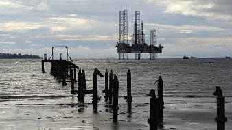 L'image présente un paysage maritime. Au premier plan, on peut voir des restes d'un vieux quai ou d'un embarcadère, avec plusieurs piliers en bois émergent de l'eau. L'eau est calme, et sa surface reflète la lumière, créant une ambiance paisible. À l'arrière-plan, une plateforme pétrolière se dresse majestueusement, avec ses grandes structures métalliques et ses grues, accentuant le contraste entre la nature et l'infrastructure industrielle. Le ciel est nuageux, laissant entrevoir une palette de gris, avec des nuances plus claires et plus sombres. L'ensemble de la scène évoque un mélange de sérénité et de robustesse, témoignant de l'interaction entre l'homme et l'environnement marine.