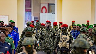 L'image représente un groupe de militaires rassemblés dans un intérieur lumineux. Au centre, un homme en uniforme se distingue par son attitude imposante. Il porte un camouflage militaire et un béret rouge. Les autres soldats autour de lui sont vêtus d'uniformes similaires, certains portant des masques de camouflage. Leurs bérets varient en couleur, notamment le rouge et le vert, créant un contraste visuel. L'ensemble évoque une atmosphère de discipline et de solidarité au sein des forces armées.