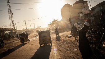 The image depicts a quiet, sandy street at sunset, where the golden light of the sun is visible on the horizon. On one side, there are simple, earth-toned walls of buildings, casting long shadows in the warm light. A few individuals are scattered along the street, with one person sitting and another standing near a donkey. The atmosphere suggests a peaceful, rural environment, with minimal traffic and a sense of tranquility as day transitions to night.