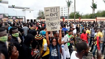 The image depicts a large crowd of people gathered in a protest setting. One individual is holding a sign that reads "KILL RAPE EXTORT END SARS NOW." The atmosphere appears to be serious and unified, with many participants wearing masks. The background includes some trees and buildings, indicating an outdoor urban environment. The protest is likely focused on social issues related to law enforcement.