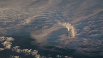 L'image présente une vue aérienne d'un paysage nuageux. On peut distinguer plusieurs cheminées d'une centrale, émettant des colonnes de fumée blanche qui se fondent dans les nuages environnants. Les nuages eux-mêmes paraissent denses et vaporeux, créant une atmosphère mystique. La lumière semble filtrer à travers le brouillard, ajoutant une touche douce au paysage. Dans l'ensemble, l'image évoque une impression de grandeur et de puissance, avec la nature et l'industrialisation coexistant dans un environnement nuageux.