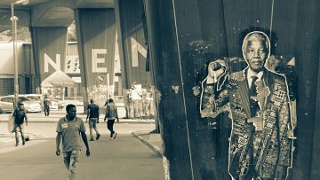 Dans cette image en noir et blanc, on voit une scène urbaine. À droite, un poster représentant Nelson Mandela est collé contre un grand pilier, le montrant avec un poing levé en signe de résistance. Le poster est texturé et présente des motifs. À gauche, un homme marche sur un chemin urbain, entouré d'autres personnes, qui semblent se diriger vers un lieu dont on aperçoit le nom inscrit en grandes lettres « NEAM » à l'arrière-plan. L'atmosphère de la scène évoque un mélange d'histoire, de mémoire et de vie quotidienne dans un environnement animé.