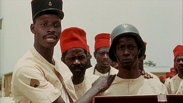 L'image montre plusieurs hommes, en uniformes, rassemblés dans un environnement clair. Au premier plan, un homme se tient à gauche, portant un chapeau noir avec un insigne et une tenue beige. Il regarde vers la droite avec une expression sérieuse. À côté de lui, un autre homme, portant un casque militaire et un uniforme beige, arbore un air grave. Derrière eux, plusieurs autres hommes regardent également dans la même direction. Tous semblent engagés dans une discussion ou une situation importante. Les couleurs dominantes de l'image sont le beige des uniformes et le rouge vif des chapeaux de certains des hommes.