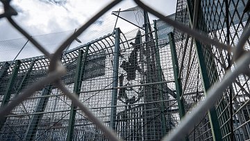 L'image représente une structure de sécurité, probablement une clôture ou un mur de protection. On aperçoit des barrières métalliques en grillage, qui sont transparentes et permettent de voir à travers. À l'intérieur, on distingue une installation, probablement en hauteur, marquée par des éléments solides tels que des poteaux. Le ciel en arrière-plan est nuageux, laissant entendre une lumière diffuse. L'ensemble dégage une impression d'isolement et de confinement, accentuée par la perspective de la clôture.