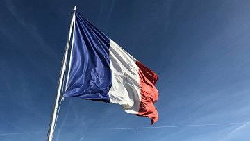 L'image montre un drapeau, qui est le drapeau tricolore français. Il est composé de trois bandes verticales : bleu à gauche, blanc au centre et rouge à droite. Le drapeau est agité par le vent, ce qui donne une impression de mouvement. À l'arrière-plan, on peut imaginer un ciel clair, probablement d'une belle journée ensoleillée, avec quelques traînées de nuages dans un bleu vif. Le contraste entre les couleurs du drapeau et la luminosité du ciel crée une image frappante et symbolique.