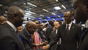 Dans cette image, nous voyons un groupe de personnes rassemblées dans un espace intérieur, probablement lors d'un événement officiel ou d'une conférence. Parmi eux se trouve un homme en costume noir avec une cravate, qui semble être un dirigeant ou une personne importante. Il tend la main pour saluer un autre homme, également en costume. Les autres personnes présentes, principalement habillées formellement, regardent avec intérêt. L'atmosphère est conviviale et respectueuse, avec des sourires et des échanges amicaux. Les murs sont décorés de manière simple, et un éclairage doux illumine la scène.