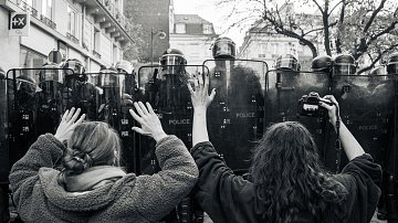 L'image montre une scène de manifestation en noir et blanc. À l'avant-plan, deux personnes se tiennent face à des policiers en tenue anti-émeute. Les manifestants, une femme brune avec les cheveux longs et une autre avec des cheveux bouclés, lèvent les bras en signe de protestation ou de désescalade. Les policiers sont rangés en formation derrière des boucliers, créant une barrière. L'environnement urbain est visible en arrière-plan, avec des bâtiments peut-être typiques de la ville. L'atmosphère est tendue, reflétant un moment de confrontation pacifique mais chargée d'émotion.