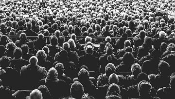 L'image montre une immense foule de personnes rassemblées dans un espace, probablement lors d'un événement ou d'un concert. Les individus sont vus de dos, et leurs têtes sont variées en termes de coiffure et de couleur de cheveux. La majorité des personnes semblent concentrées et immergées dans l'événement, créant une atmosphère collective. Les jeux d'ombres et de lumières donnent à l'image un aspect noir et blanc, accentuant la densité de la foule et le sentiment d'unité parmi les spectateurs. On peut presque sentir l'énergie vibrante qui se dégage de ce groupe immense.