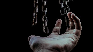 L'image montre une main tendue vers le haut, en direction de chaînes suspendues qui descendent. La main, légèrement ouverte, semble chercher à atteindre les chaînes, qui sont bien visibles sur un fond sombre. Les chaînes sont métalliques, avec une texture rustique, et elles évoquent une sensation d'emprisonnement ou de lutte. L'éclairage met en valeur les détails des chaînes et de la main, créant une atmosphère à la fois troublante et poignante. Cette composition suggère un thème de quête ou de désir de liberté.