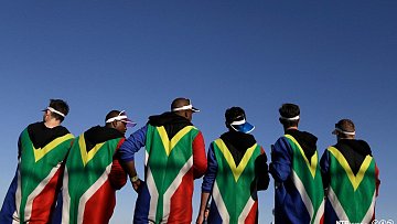 L'image montre un groupe de personnes de dos, habillées dans des capes aux couleurs du drapeau sud-africain, dominant les tons vert, noir, jaune et rouge. Leurs silhouettes sont nettement visibles contre un ciel bleu clair. Les individus semblent détendus et peuvent être en train de profiter d'un événement ou d'une célébration. Certains portent des visières sur la tête, ajoutant une touche festive à leur tenue. L'ensemble dégage une ambiance dynamique et communautaire, soulignant la fierté et l'identité culturelle.