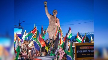 L'image montre une scène de manifestation en plein air, sous un ciel bleu. Au centre, se dresse une grande statue de Nelson Mandela, levant un poing en signe de force et de détermination. Autour de la statue, des manifestants tiennent une multitude de drapeaux, notamment des drapeaux sud-africains et palestiniens, créant un mélange de couleurs vives. Des pancartes sont visibles, certaines appelant à un cessez-le-feu et dénonçant des injustices. Les visages des participants expriment à la fois la passion et l'engagement pour leur cause. L'atmosphère générale est celle de la solidarité et de la lutte pour les droits humains.
