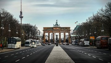 Cette image représente un point de vue d'une grande avenue à Berlin, avec en toile de fond la célèbre porte de Brandebourg. Cette porte emblématique, ornée d'une sculpture représentant un char tiré par des chevaux, est encadrée par des arbres dénudés, typiques des saisons froides. À gauche, on aperçoit la tour de télévision de Berlin, un autre symbole de la ville, se dressant majestueusement dans le ciel. L'avenue est bordée de bus et de véhicules, et la chaussée est pavée de dalles claires, créant un contraste agréable avec le bitume. Le ciel est légèrement nuageux, ajoutant une ambiance calme et réfléchie à cette scène urbaine.