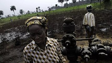 L'image montre un paysage rural où se trouvent des plantations de palmiers en arrière-plan. Au premier plan, une femme d'âge mûr, vêtue d'une blouse rayée et portant un foulard coloré, se tient près d'une infrastructure de forage pétrolier. Son expression est sérieuse et préoccupée. À l'arrière-plan, une autre personne, également en tenue décontractée, semble s'occuper de l'équipement. Le sol est noirci, suggérant une dégradation environnementale. L'atmosphère générale évoque une lutte pour préserver leur lien avec la terre face aux impacts de l'exploitation pétrolière.