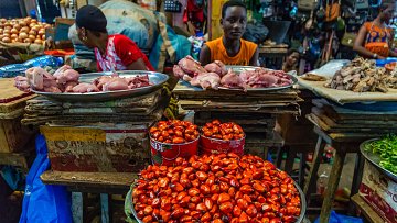 L'image montre un marché animé, où des étals en bois sont chargés de différents aliments. Au premier plan, un grand plat en métal contient des petites noix rouge vif, à côté d'autres aliments. À l'arrière-plan, des morceaux de viande sont disposés sur des plateaux, et on peut voir des vendeurs et des clients s'affairer autour. Les couleurs sont vives, avec des nuances de rouge, de marron et de vert. L'atmosphère est dynamique, remplie de sons de discussions et d'échanges.