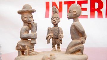 L'image présente une sculpture en bois représentant trois figures humaines stylisées. Au centre, une figure masculine, aux traits marqués, tient un objet dans les mains, probablement un outil ou un symbole culturel. Il est entouré de deux autres sculptures, l'une à gauche, qui porte un chapeau large, et l'autre à droite, avec une coiffure distinctive. Les personnages semblent engagés dans une conversation ou un échange, ce qui suggère une interaction sociale. Les détails des sculptures, comme les expressions des visages et les postures, témoignent d'un art tribal riche en histoire et en culture. Le fond de l'image est flou, mettant l'accent sur les sculptures elles-mêmes.