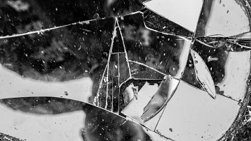 L'image présente un miroir brisé, dont les morceaux reflètent de manière fragmentée l'environnement. Les éclats de verre montrent des bords abrupts et irréguliers, créant une texture variée. La lumière joue sur la surface des fragments, produisant des éclats scintillants. La composition monochrome en noir et blanc accentue les contrastes entre les zones sombres et lumineuses, apportant une ambiance dramatique à l'ensemble. L'impression générale évoque une sensation de fragilité et de désordre, tout en incitant à la réflexion sur les thèmes de la rupture et de la beauté dans l'imparfait.
