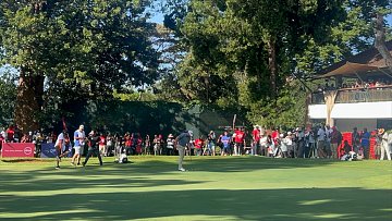 Imaginez un vaste terrain de golf, baigné par la lumière du soleil. Au premier plan, deux joueurs se concentrent sur le putting green, faisant preuve de délicatesse et de précision. L'un d'eux est en position de putt, tandis que l'autre observe attentivement. À l'arrière-plan, des spectateurs en tenue décontractée, vêtus de couleurs variées, attendent avec excitation, certains filmant ou prenant des photos. Autour du green, de grands arbres fournissent de l'ombre, ajoutant à la beauté de l'environnement. À droite, on aperçoit un bâtiment avec des balcons, où d'autres personnes sont rassemblées pour profiter du spectacle. L'atmosphère est vivante, remplie de murmures, de rires et de l'anticipation d'un moment décisif dans le jeu.