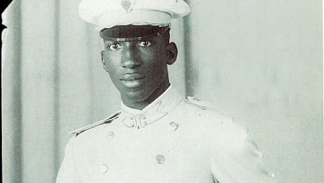 L'image montre un jeune homme en tenue militaire. Il porte un uniforme blanc qui se compose d'une chemise et d'un pantalon assortis, avec une ceinture qui souligne sa taille. Sur sa tête, il a une casquette blanche ornée d'un insigne. Son expression est sérieuse et déterminée. L'arrière-plan est neutre, probablement un drap ou un fond uni, ce qui met en valeur sa silhouette et son uniforme.