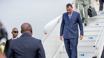 L'image montre un homme qui descend des marches d'un avion. Il porte un costume bleu foncé et des lunettes de soleil. Son expression semble concentrée tandis qu'il s'avance avec assurance. À côté de lui, un autre homme en costume noir observe attentivement. En arrière-plan, on aperçoit des membres du personnel de l'aéroport, ainsi que l'avion stationné. L'atmosphère peut sembler formelle et dynamique, suggérant un moment important, vraisemblablement une arrivée ou une sortie officielle.