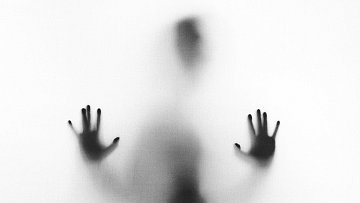 L'image représente une silhouette floue d'une personne, en arrière-plan léger. La figure se trouve proche d'une surface translucide, créant un effet de brouillard. On peut distinguer des mains, les paumes dirigées vers l'avant, comme si la personne essayait de pousser ou de traverser cette barrière. Les contours sont indistincts, évoquant une sensation de mystère et d'isolement. L'ambiance générale est à la fois intrigante et un peu mélancolique.