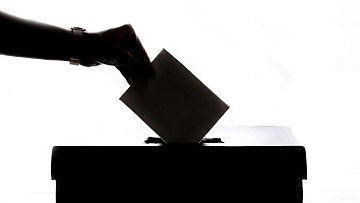 L'image représente une scène de vote. On peut voir une main tendue vers une urne, prête à y insérer un bulletin de vote. La silhouette de la main et de l'urne est visible contre un fond lumineux, créant un contraste fort. L'urne est de forme rectangulaire et se trouve sur une surface plane. Cette image évoque le processus démocratique et l'importance de l'engagement civique.