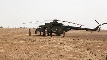 L'image montre un hélicoptère posé sur un terrain désertique, avec un sol caillouteux et sec. Autour de l'hélicoptère, plusieurs personnes sont rassemblées, certaines en uniforme militaire. La scène semble se dérouler dans une région aride, avec peu de végétation en arrière-plan.